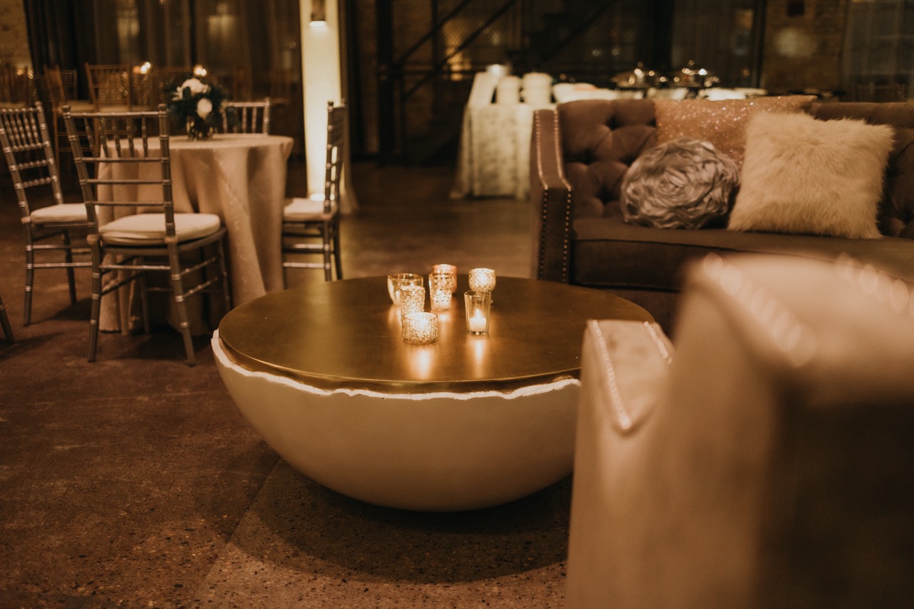 A warm, atmospheric wedding lounge area featuring a cluster of lit tea light candles in clear glass votive holders on a round gold coffee table in Minnesota.