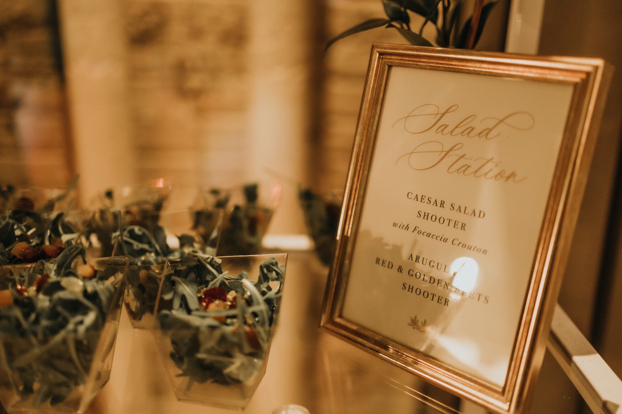 Elegant gold picture frame displaying a wedding salad station menu next to small glass cups filled with fresh arugula salads for an event in Minnesota.