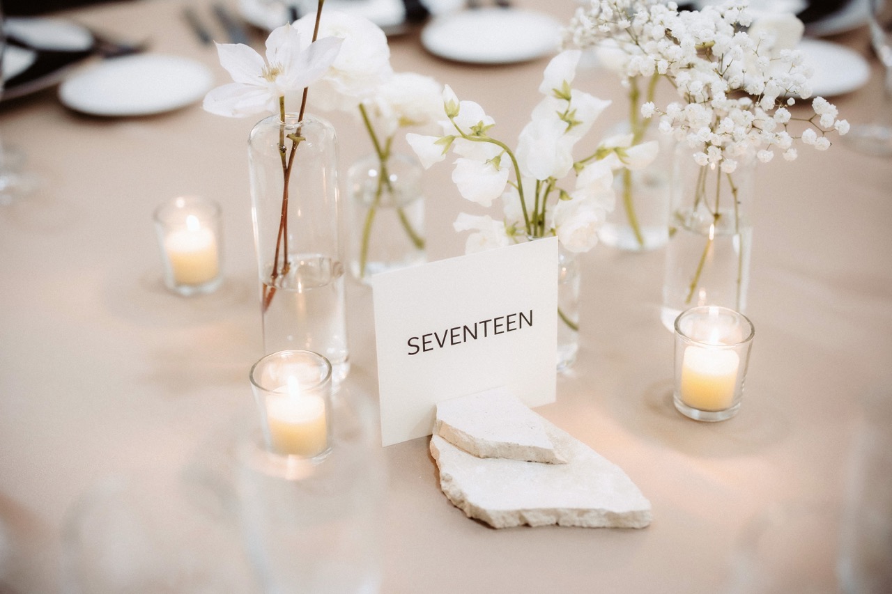 Minimalist modern wedding table number seventeen styled with flat stone bases, glass bud vases with white flowers, and clear glass votive holders with lit tea light candles.