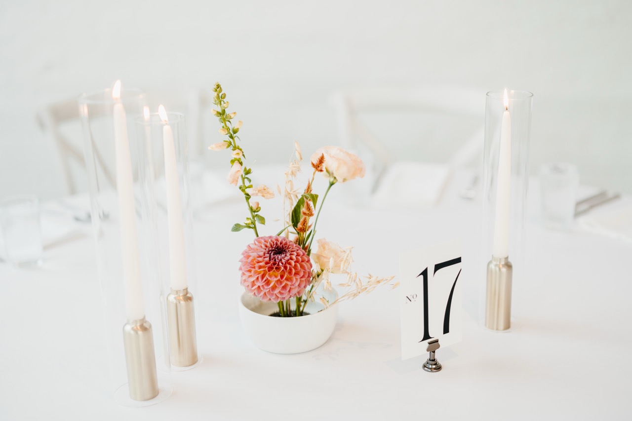 Modern wedding table number seventeen card in a small metal holder on a white reception table with a dahlia centerpiece and tall taper candles in glass chimneys.