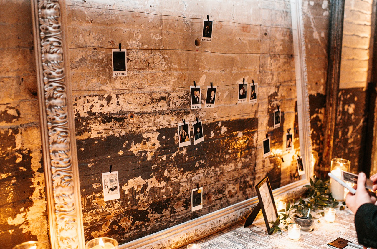 Interactive wedding guest book display featuring Polaroid film photos clipped to wires inside a large ornate silver frame against a rustic brick wall in Minnesota.