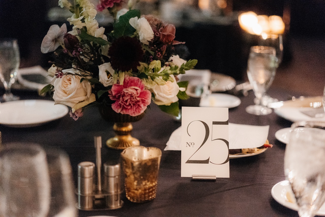 Modern elegant wedding table number twenty-five on a dark reception table with a gold floral centerpiece and votive candles.