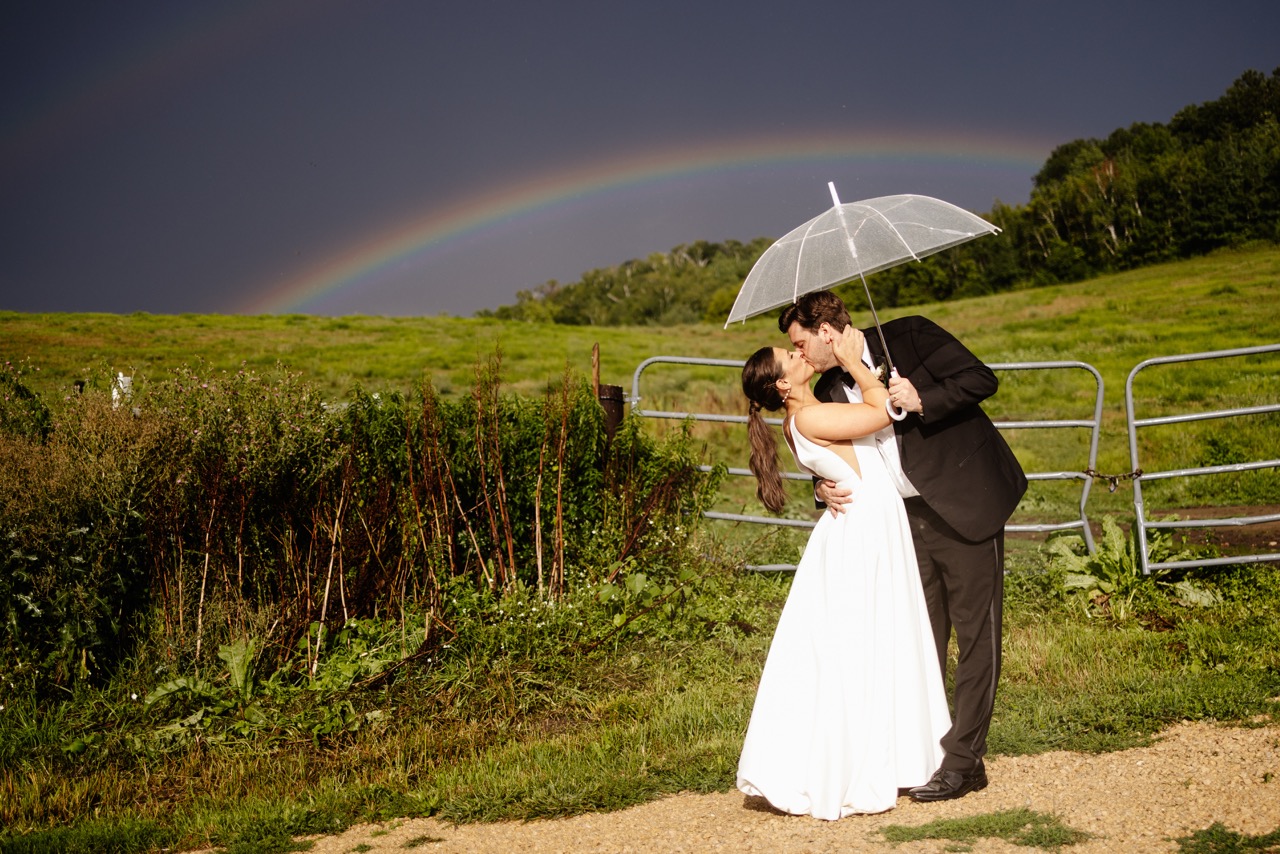 Bride and groom kissing under a clear wedding umbrella in a green field with a vibrant rainbow and dark storm sky in Minnesota.