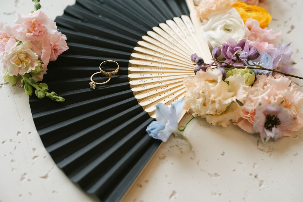 Gold wedding rings resting on a pleated black paper hand fan with bamboo spokes, surrounded by pastel wedding flowers for a Minnesota event.
